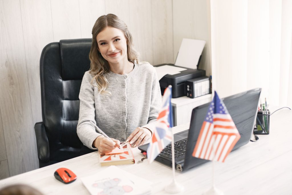 Mulher sorridente em uma mesa com bandeiras britânica e americana, transmitindo a importância do inglês no compliance e ESG.