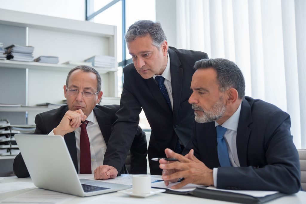 Três homens de terno lendo documentos de compliance e ESG em inglês em um notebook dentro de um escritório.