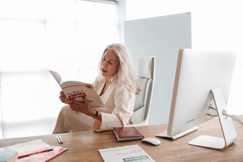 Mulher de terno branco, sentada em um escritório, lendo um livro de inglês para lideranças.